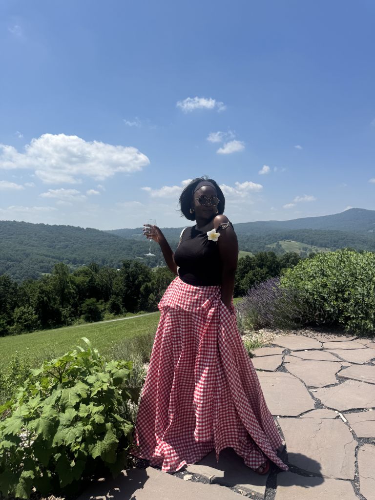 Minnie in a sweeping red-and-white gingham maxi skirt she made by hand, standing against a sunlit mountain view
